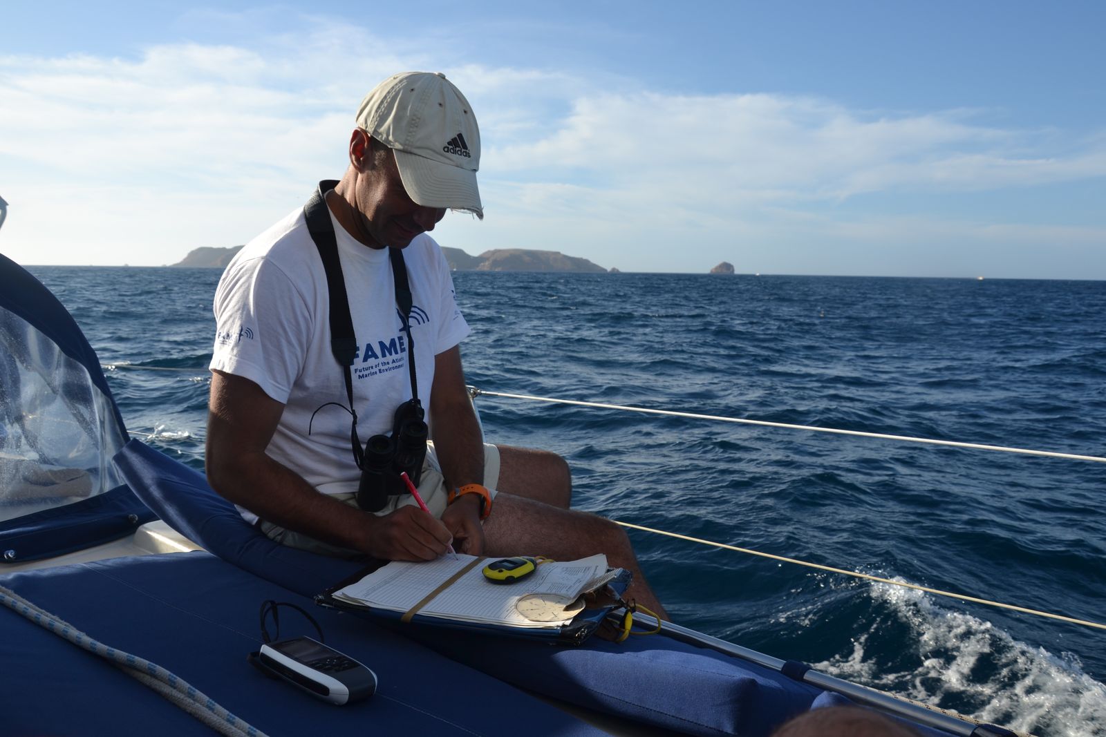 Investigador a bordo, com binóculos ao pescoço, a registar observações no âmbito do censo ESAS. Ao fundo, observa-se a ilha da Berlenga e, no barco, parte do equipamento utilizado no censo, incluindo um cronómetro e um GPS.